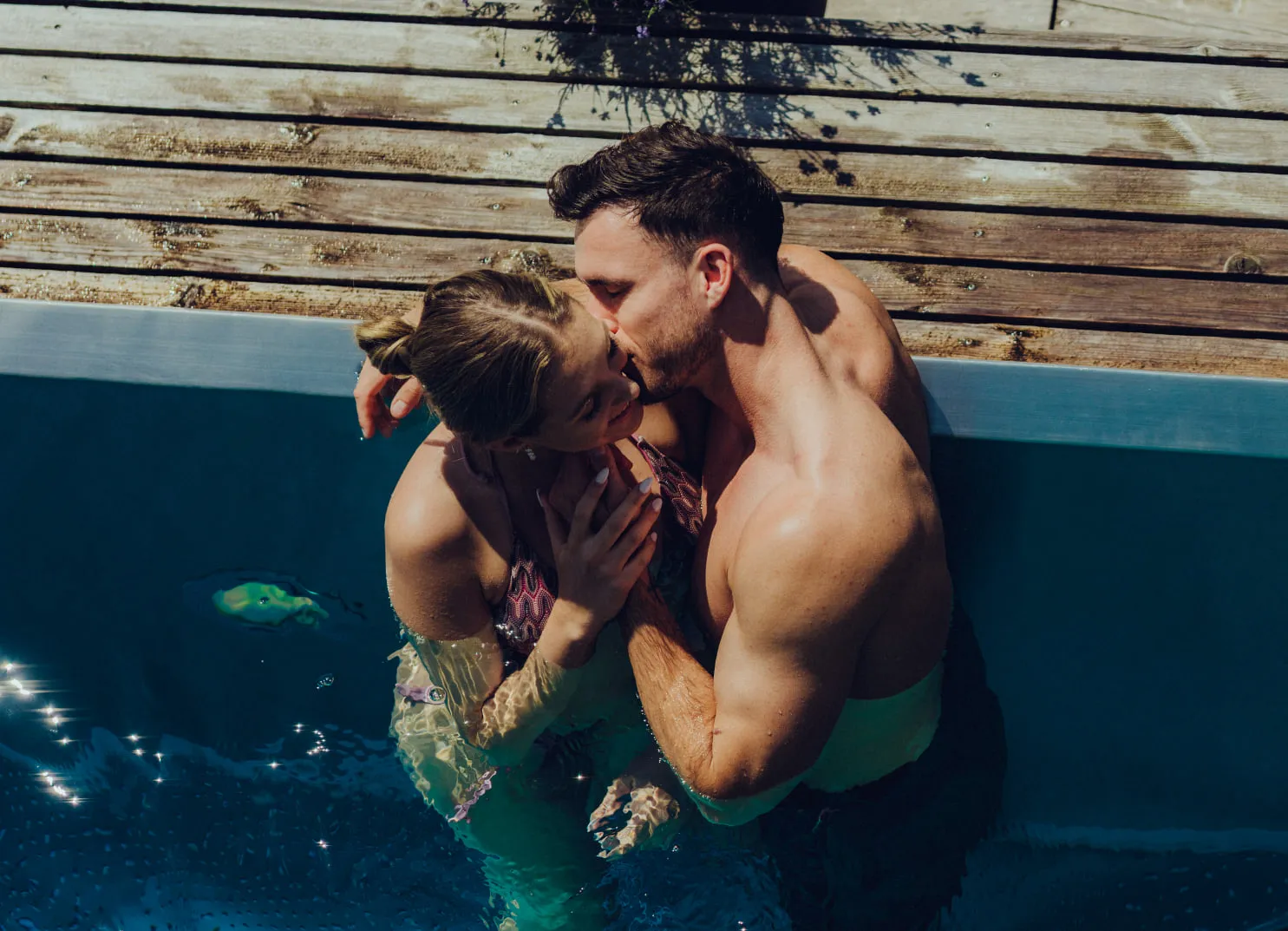 Paar kuschelt im Pool, umgeben von Holzdeck.